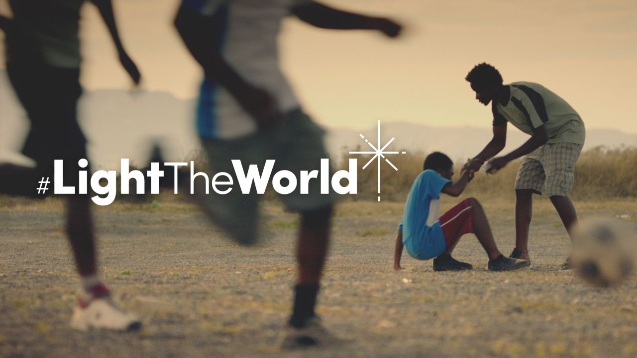 Un niño ayuda a otro niño a levantarse después de haberse caído jugando al fútbol (del video Ilumina el Mundo)